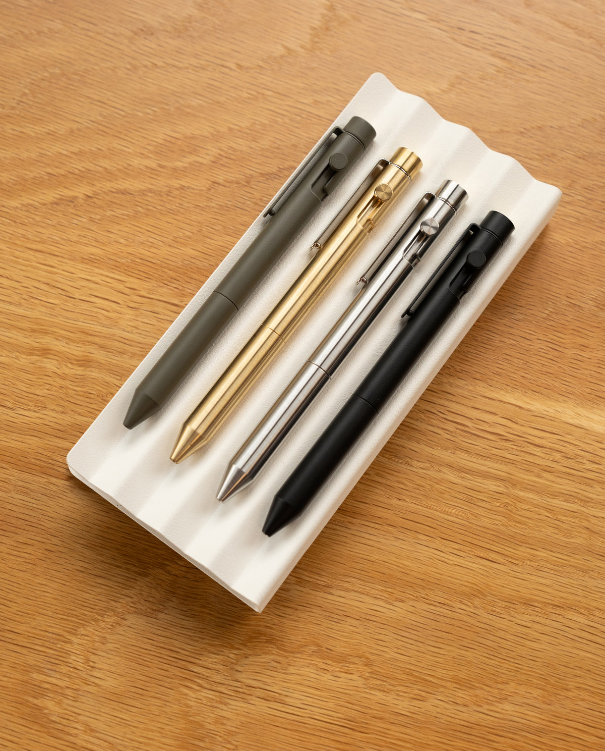 Four INVENTERY Bolt Action Pens in onyx are neatly arranged on a white, grooved tray placed on a wooden surface. Each steel pen features a durable Cerakote ceramic coating for added resilience and style.