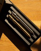 Two INVENTERY Bolt Action Pens—one crafted from solid brass with a gold finish and the other adorned with a sleek silver Cerakote ceramic coating—are displayed side by side in a black case atop a wooden surface, sunlight casting elongated shadows across the scene.