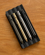 Four INVENTERY Bolt Action Pens (Stainless) in matte black, gold, silver, and black are arranged on a ridged black tray atop a wooden surface—each pen delivering style and lasting durability for daily writing tasks.