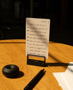 A handwritten to-do list on a small notepad sits upright on a wooden table, next to a pen, an HMM Clips (Set of 2 - Black), a round black object, and part of another notepad, all illuminated by sunlight.