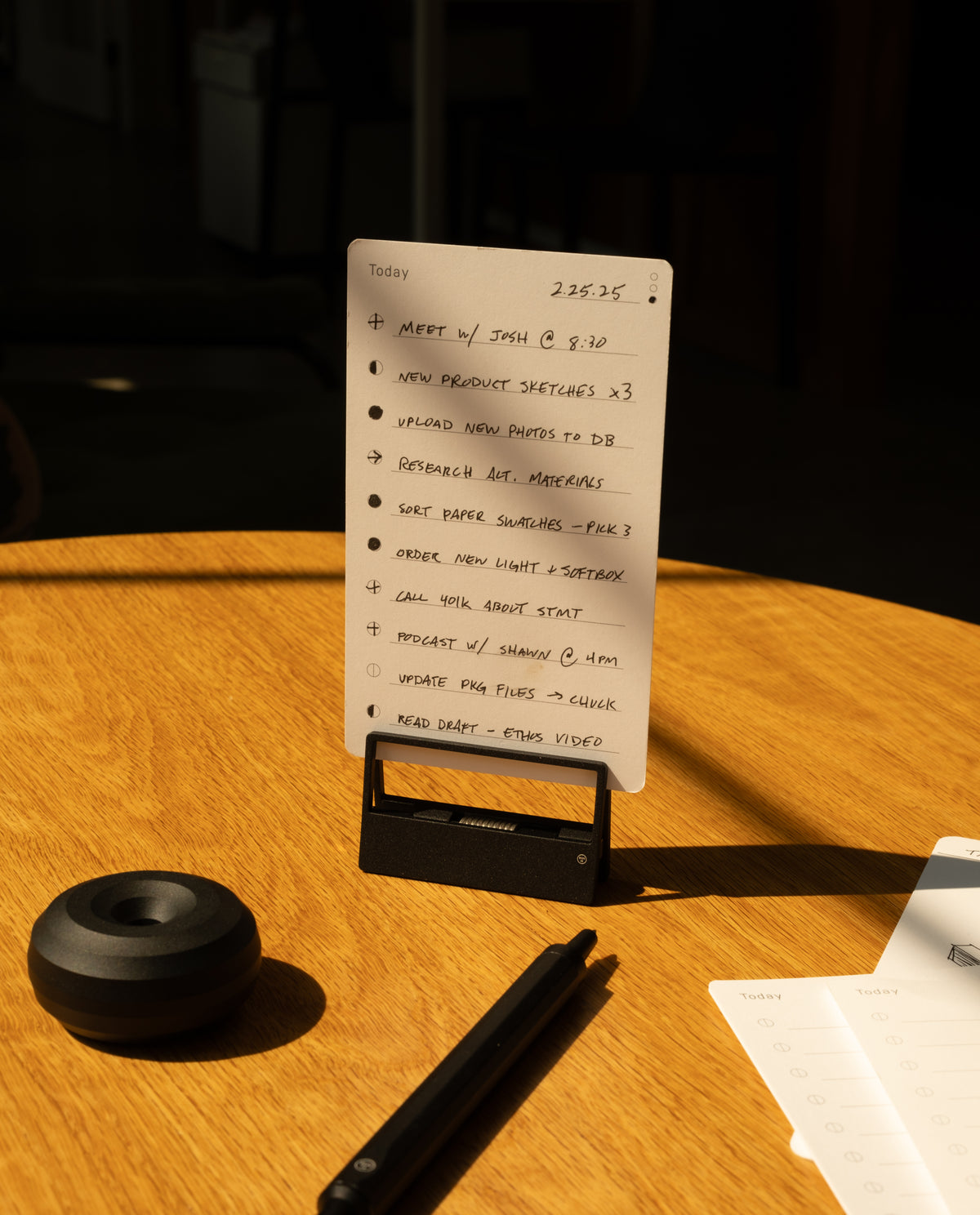 A handwritten to-do list on a small notepad sits upright on a wooden table, next to a pen, an HMM Clips (Set of 2 - Black), a round black object, and part of another notepad, all illuminated by sunlight.
