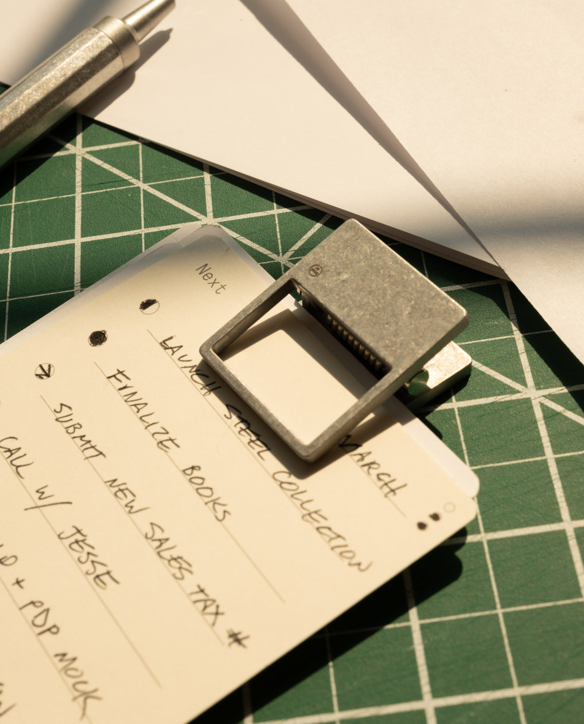 A close-up of a handwritten to-do list secured with HMM Clips (Set of 2 - Raw) to a notepad on a green cutting mat, with a pen and white paper sheets arranged neatly and sunlight illuminating the organized workspace.