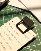 A handwritten to-do list is clipped with an HMM Clip (Set of 2 - Black) on a green cutting mat. Sunlight creates shadows, drawing attention to the list and this stylish desk organizer accessory.