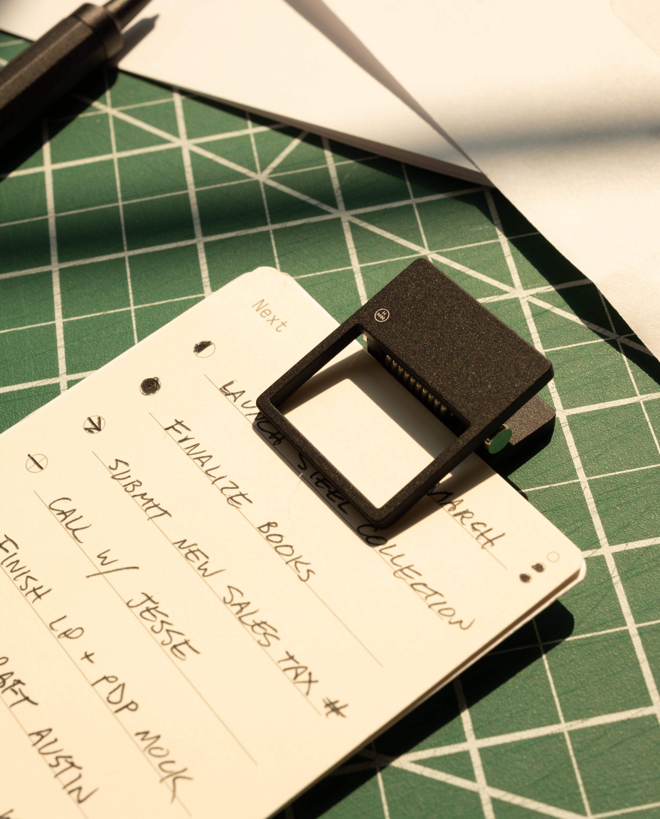 A handwritten to-do list is clipped with an HMM Clip (Set of 2 - Black) on a green cutting mat. Sunlight creates shadows, drawing attention to the list and this stylish desk organizer accessory.