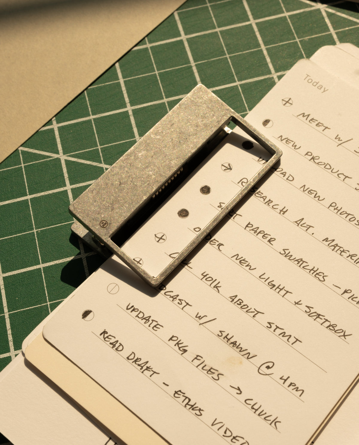 A stack of handwritten to-do lists—meetings, research, updates—is secured by HMM Clips (Set of 2 - Raw) on a green cutting mat as warm sunlight casts gentle shadows across the scene.