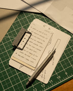 An HMM Clip (Set of 2 - Raw) secures a stack of index cards with handwritten notes and sketches, topped by a metal pen. The cards rest on a green cutting mat with grid lines, surrounded by sheets of paper.
