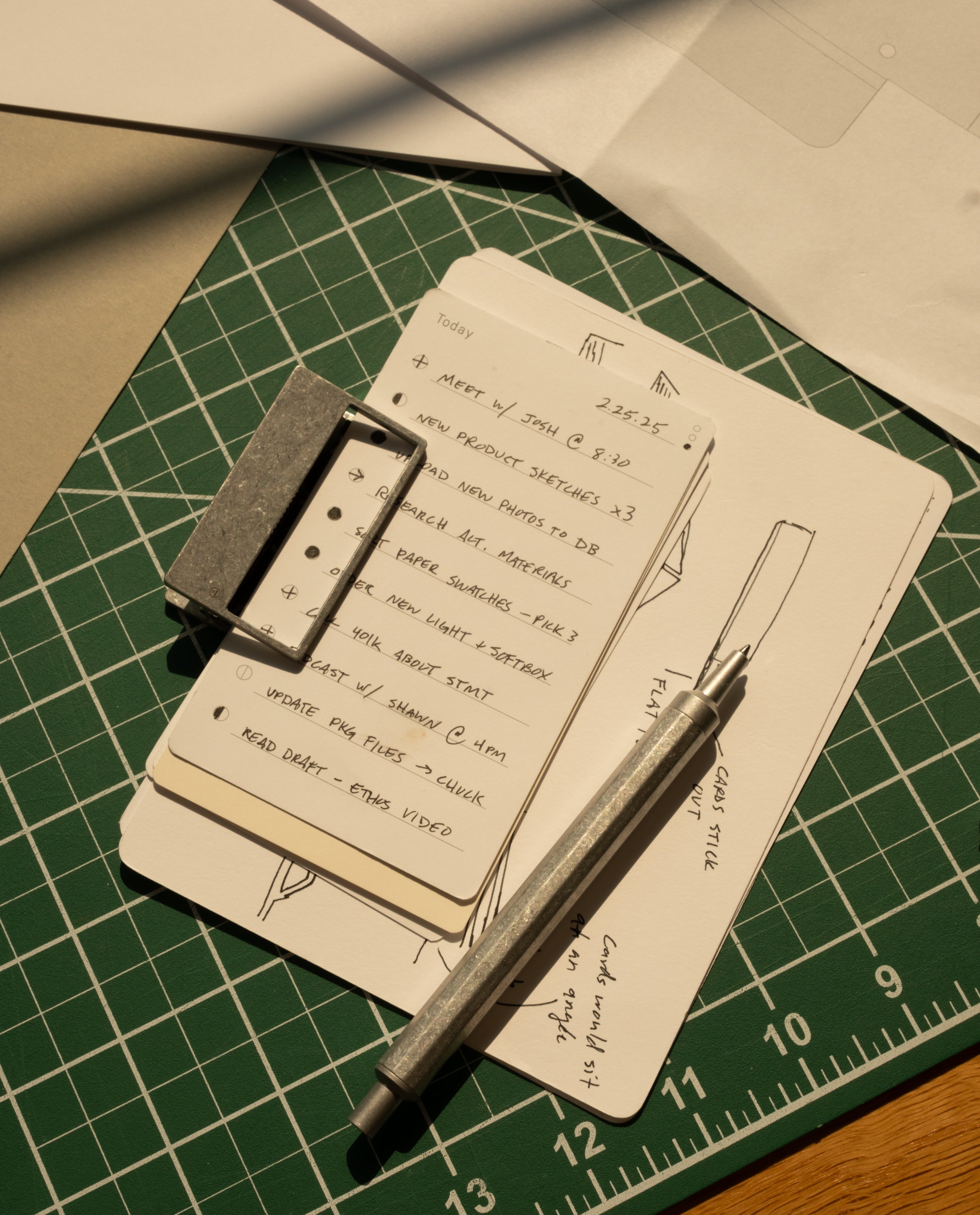 An HMM Clip (Set of 2 - Raw) secures a stack of index cards with handwritten notes and sketches, topped by a metal pen. The cards rest on a green cutting mat with grid lines, surrounded by sheets of paper.