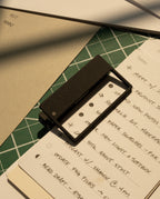 A rectangular magnifier sits on a handwritten to-do list over green and beige papers, with HMM Clips (Set of 2 - Black) and sunlight casting shadows across the organized workspace.