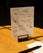 A sheet of paper, secured with HMM Clips (Set of 2 - Black), displays sketches and notes about table risers on a stand. Another sheet with handwritten notes and a black pen rests on a warmly lit wooden table.