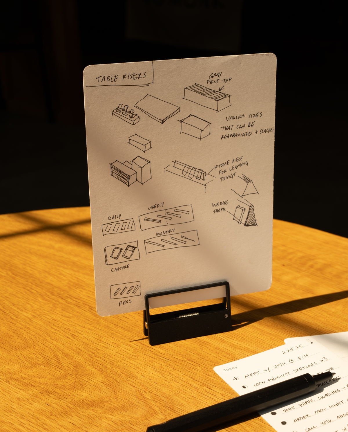 A sheet of paper, secured with HMM Clips (Set of 2 - Black), displays sketches and notes about table risers on a stand. Another sheet with handwritten notes and a black pen rests on a warmly lit wooden table.