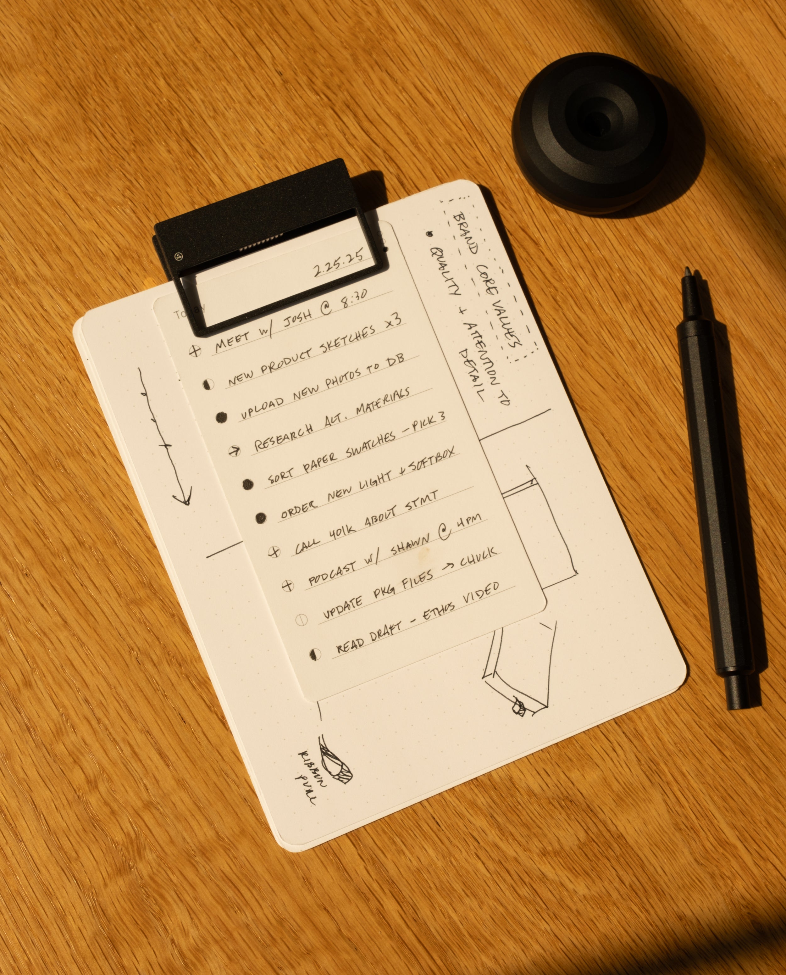 A small clipboard with a dotted notepad displays a to-do list and sketches in black ink. Nearby, HMM Clips (Set of 2 - Black) and a black pen rest on the wooden surface, sunlight casting gentle shadows across the scene.