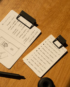 Two notepads with HMM Clips (Set of 2 - Black) lie on a wooden table—one displays a hand-drawn floor plan and notes, while the other lists tasks. A black pen, round object, and warm lighting create an organized workspace atmosphere.