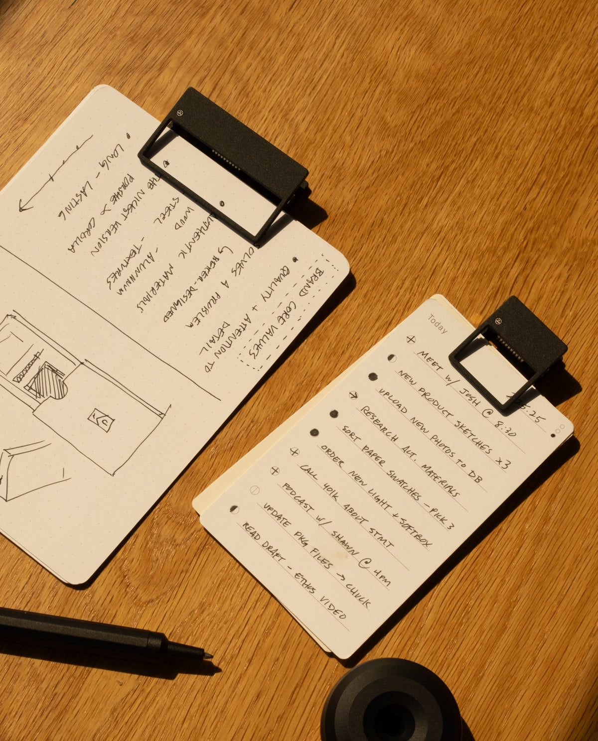 Two notepads with HMM Clips (Set of 2 - Black) lie on a wooden table—one displays a hand-drawn floor plan and notes, while the other lists tasks. A black pen, round object, and warm lighting create an organized workspace atmosphere.