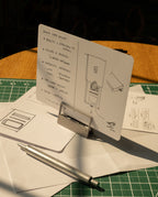 A product design sketch on a notecard rests on an HMM Clips (Set of 2 - Raw) aluminum holder, surrounded by pens, sketches, and a green cutting mat—all bathed in warm sunlight.
