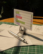 A small Gantt chart card stands on a metal holder, with scattered papers secured by HMM Clips (Set of 2 - Raw) and a silver pen on a green cutting mat, all bathed in warm sunlight.
