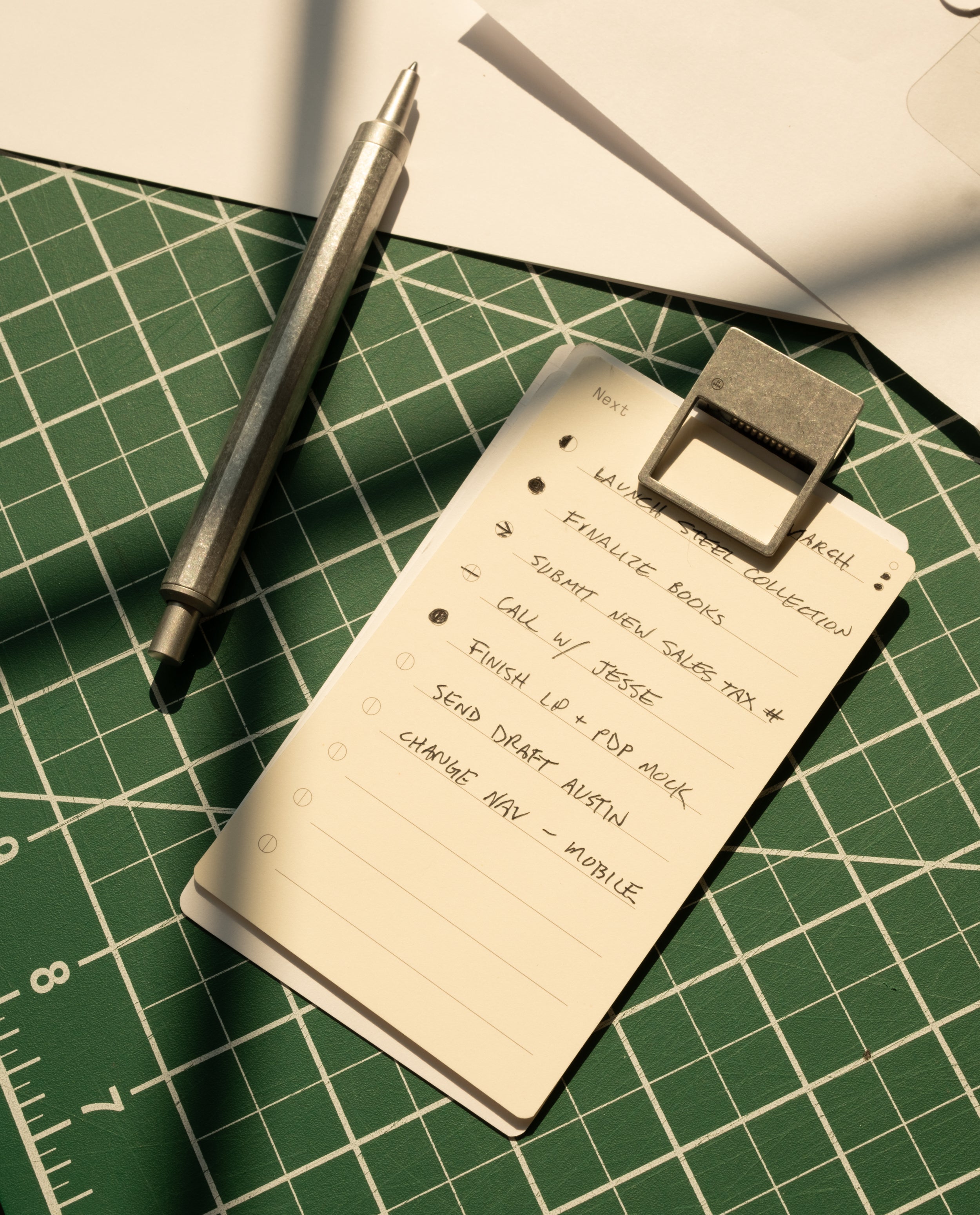 A metal pen and HMM Clips (Set of 2 - Raw) secure a handwritten to-do list on a green cutting mat, with tasks like finalizing a book collection, submitting sales tax, making calls, and sending a draft.