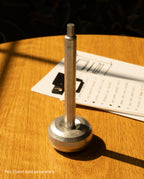 A HMM Ballpoint Pen (Raw) stands upright in a round metal base on a wooden table. In the background, there is a clipboard with paper. Text at the bottom left reads, Pen Stand sold separately.