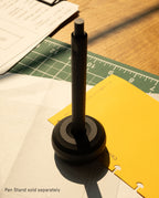 A pen stands upright in the HMM Paperweight Pen Stand (Black), a sleek desk accessory, on a desk with paper, a yellow folder, and a cutting mat. Sunlight casts shadows across the workspace. Text reads, Pen Stand sold separately.