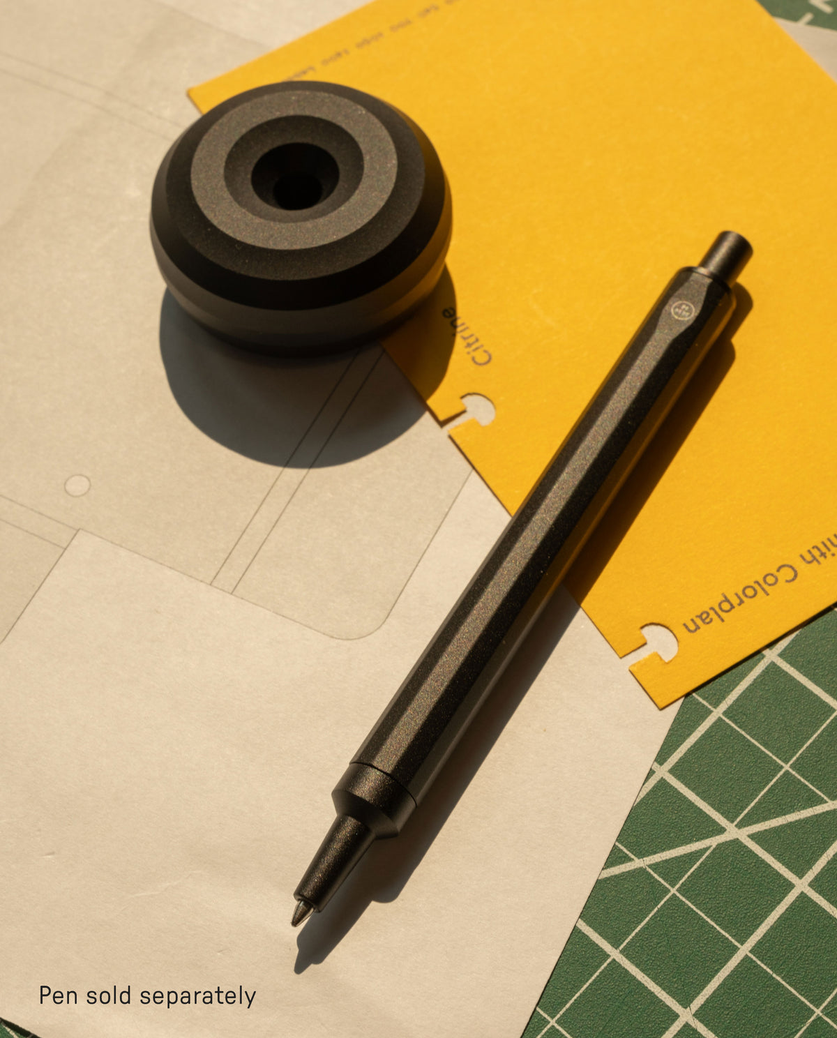 A black HMM Paperweight Pen Stand is shown on paper and a yellow folder, with sunlight casting shadows. Text in the corner reads: Pen sold separately—an ideal desk accessory for any workspace.