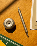 A HMM Ballpoint Pen (Raw) and a round metal object rest on a wooden desk near a ruler, color swatches, and a folder, all brightly illuminated by sunlight.