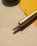 The HMM Ballpoint Pen (Raw) rests on sheets of paper—one yellow with holes and another grey with faint technical drawings—under warm light.