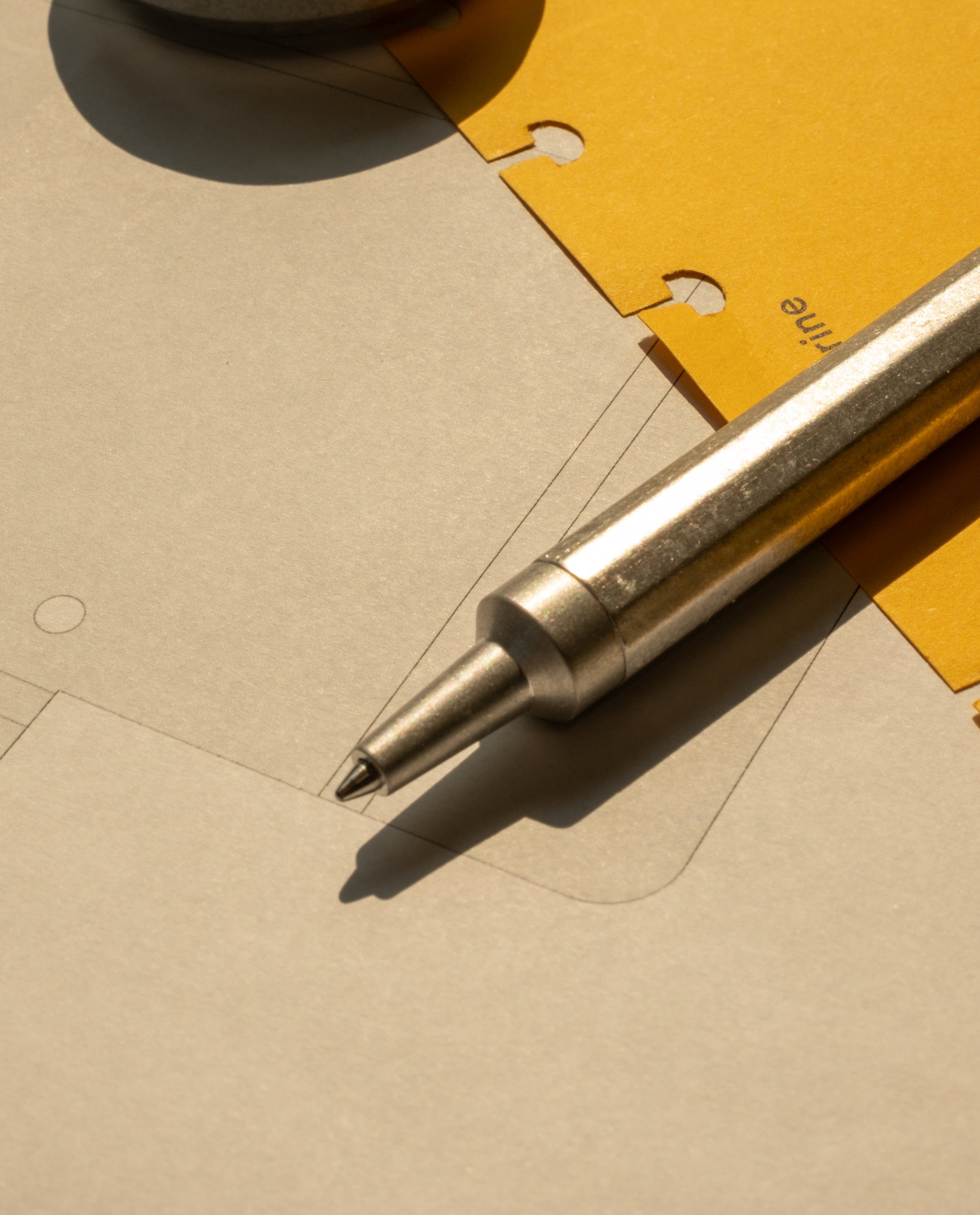 The HMM Ballpoint Pen (Raw) rests on sheets of paper—one yellow with holes and another grey with faint technical drawings—under warm light.