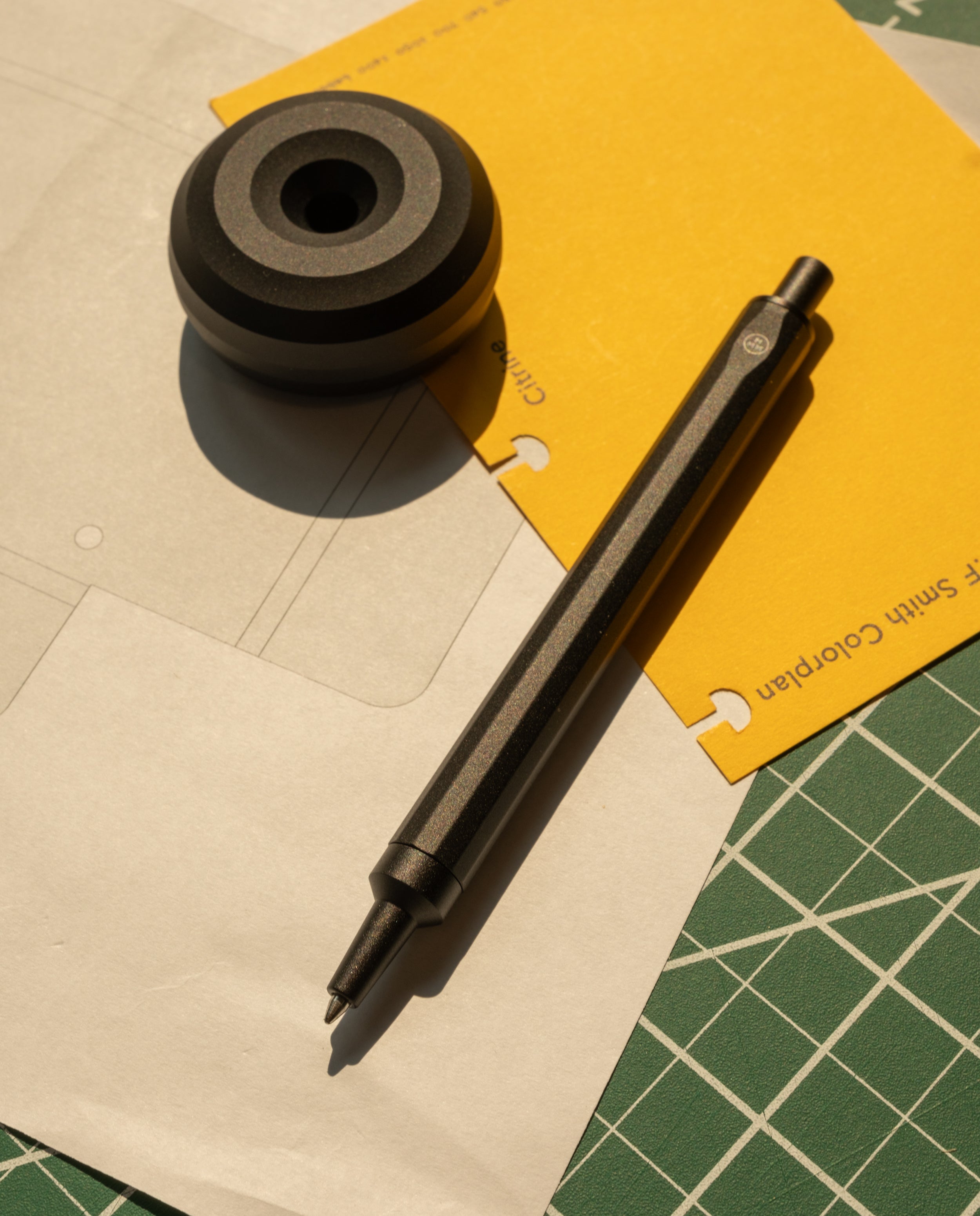 The HMM Ballpoint Pen (Black) and a round pen holder rest on stacked yellow and white papers atop a green cutting mat in bright sunlight.