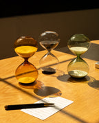 A 15 Minute Hourglass with green sand sits on a wooden table, sunlight casting shadows across the surface and highlighting the analog timer. A pen and a small notepad with writing are also on the table.