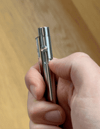 A hand holds the INVENTERY Bolt Action Pen (Stainless), thumb pressing the top button. The blurred wooden background accentuates its sleek, stainless steel design.