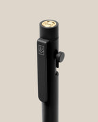 A close-up of the INVENTERY Bolt Action Pen (Onyx), a sleek black pen made from steel with a luxurious Cerakote ceramic coating. It features a gold-colored button on top, a side clip engraved with a geometric logo, and a small round side button, all set against a beige background.