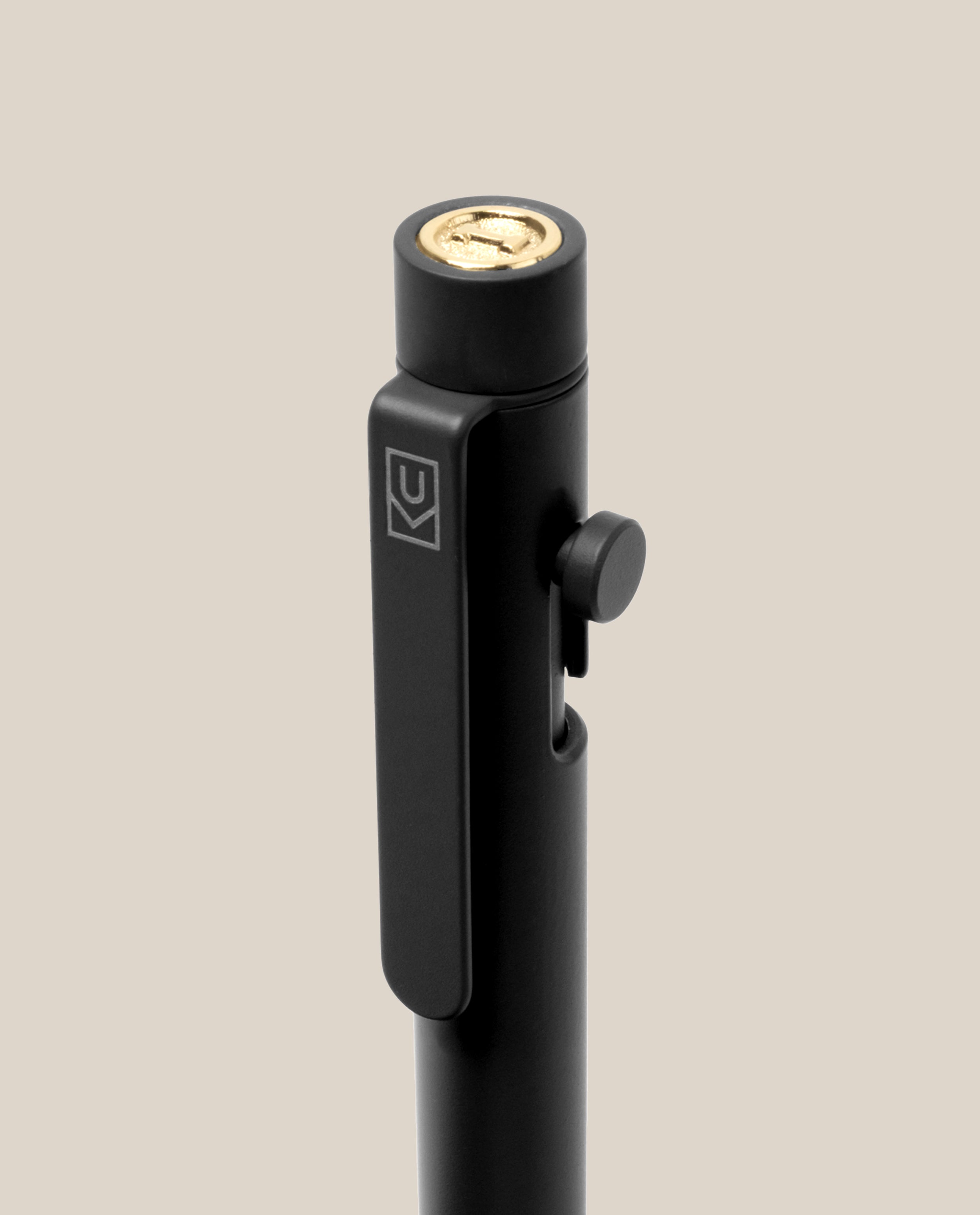 A close-up of the INVENTERY Bolt Action Pen (Onyx), a sleek black pen made from steel with a luxurious Cerakote ceramic coating. It features a gold-colored button on top, a side clip engraved with a geometric logo, and a small round side button, all set against a beige background.