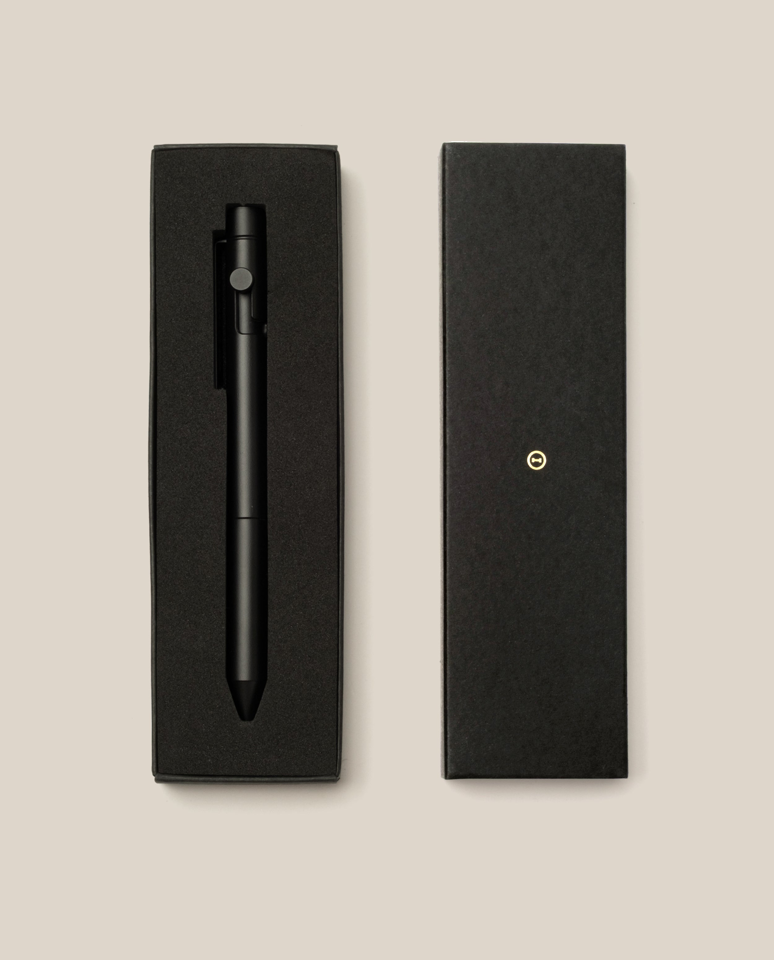 An INVENTERY Bolt Action Pen (Onyx) in matte black, finished with a durable Cerakote ceramic coating for enhanced sophistication and resilience, rests elegantly in a rectangular black gift box lined with foam padding. The box lid is placed beside it on a beige background.