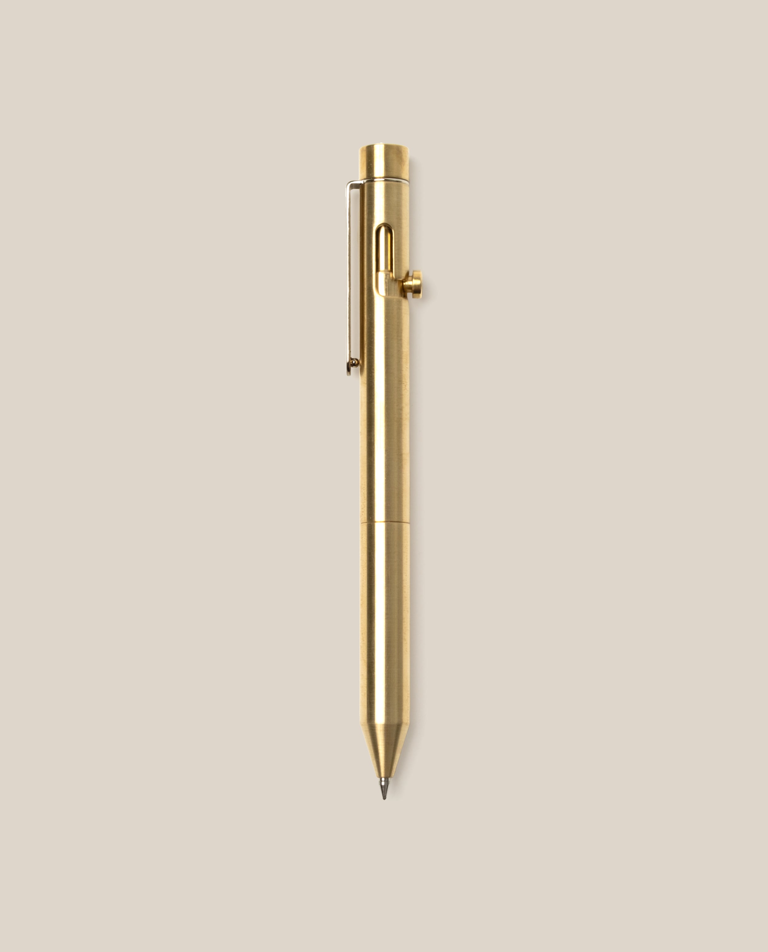 The INVENTERY Bolt Action Pen (Brass), featuring a sleek clip and a smooth bolt-action mechanism, is centered vertically on a plain beige background.