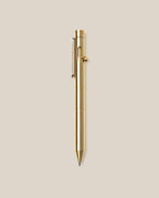 The INVENTERY Bolt Action Pen (Brass), featuring a sleek clip and a smooth bolt-action mechanism, is centered vertically on a plain beige background.