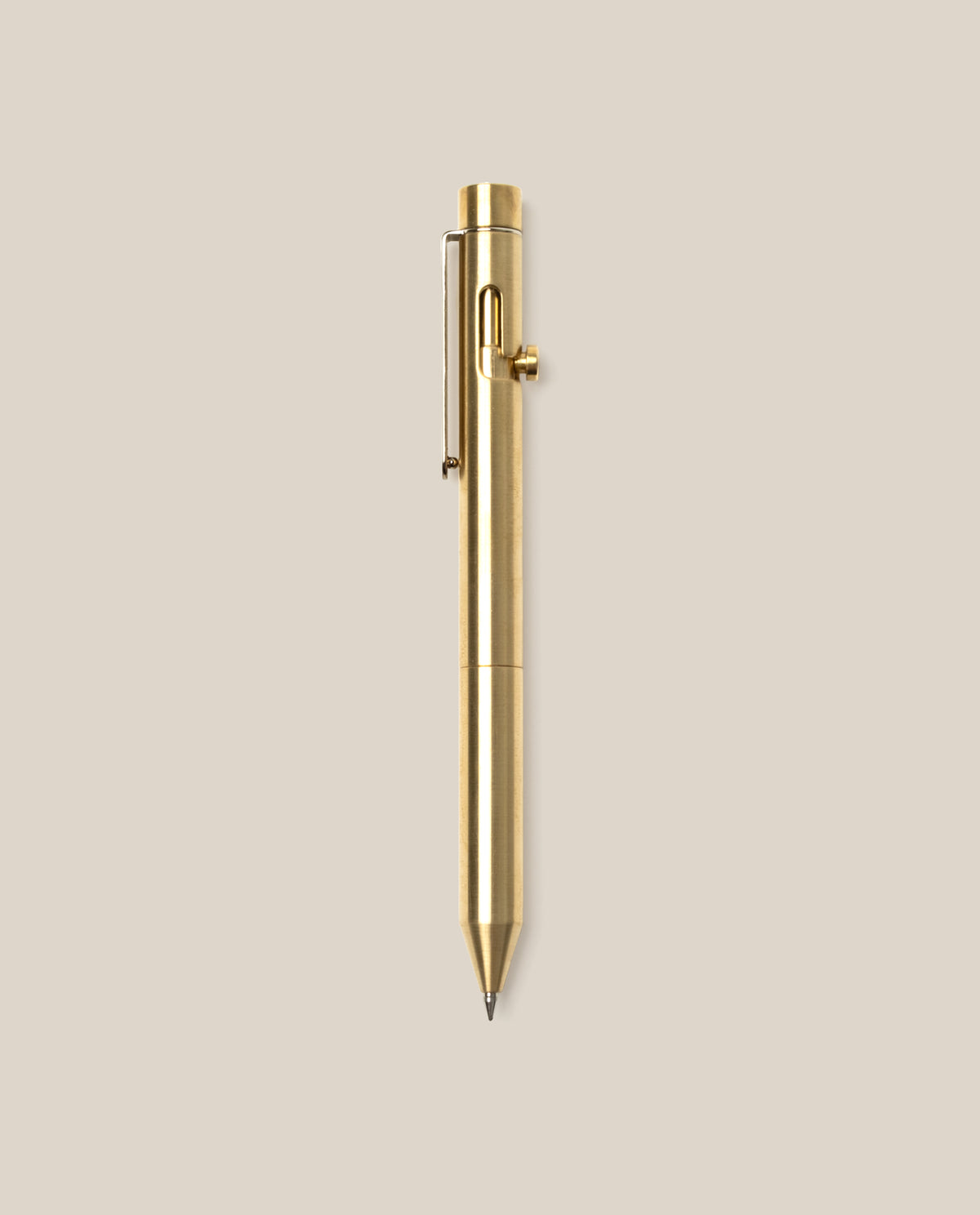 The INVENTERY Bolt Action Pen (Brass), featuring a sleek clip and a smooth bolt-action mechanism, is centered vertically on a plain beige background.