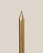 A close-up of the INVENTERY Bolt Action Pen (Brass), crafted from solid brass with a pointed tip and finished in durable Cerakote ceramic coating, positioned vertically against a plain, light beige background.