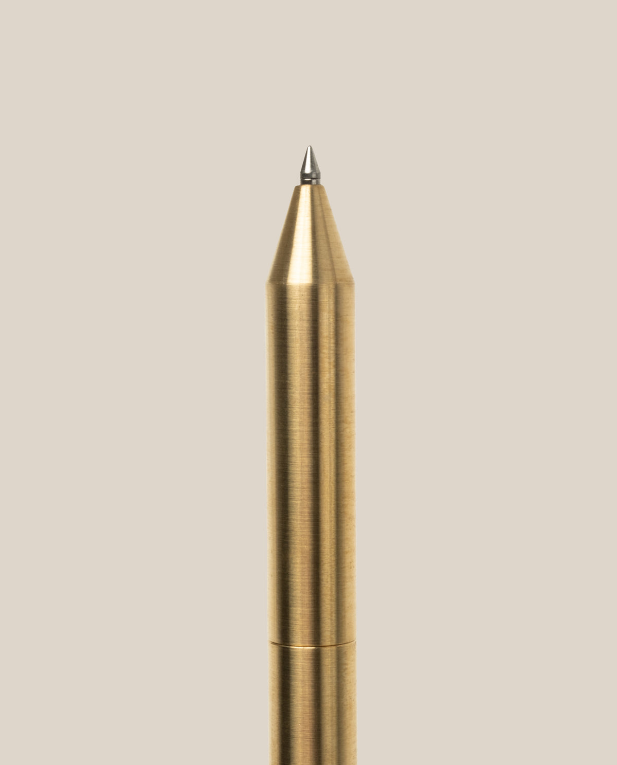 A close-up of the INVENTERY Bolt Action Pen (Brass), crafted from solid brass with a pointed tip and finished in durable Cerakote ceramic coating, positioned vertically against a plain, light beige background.