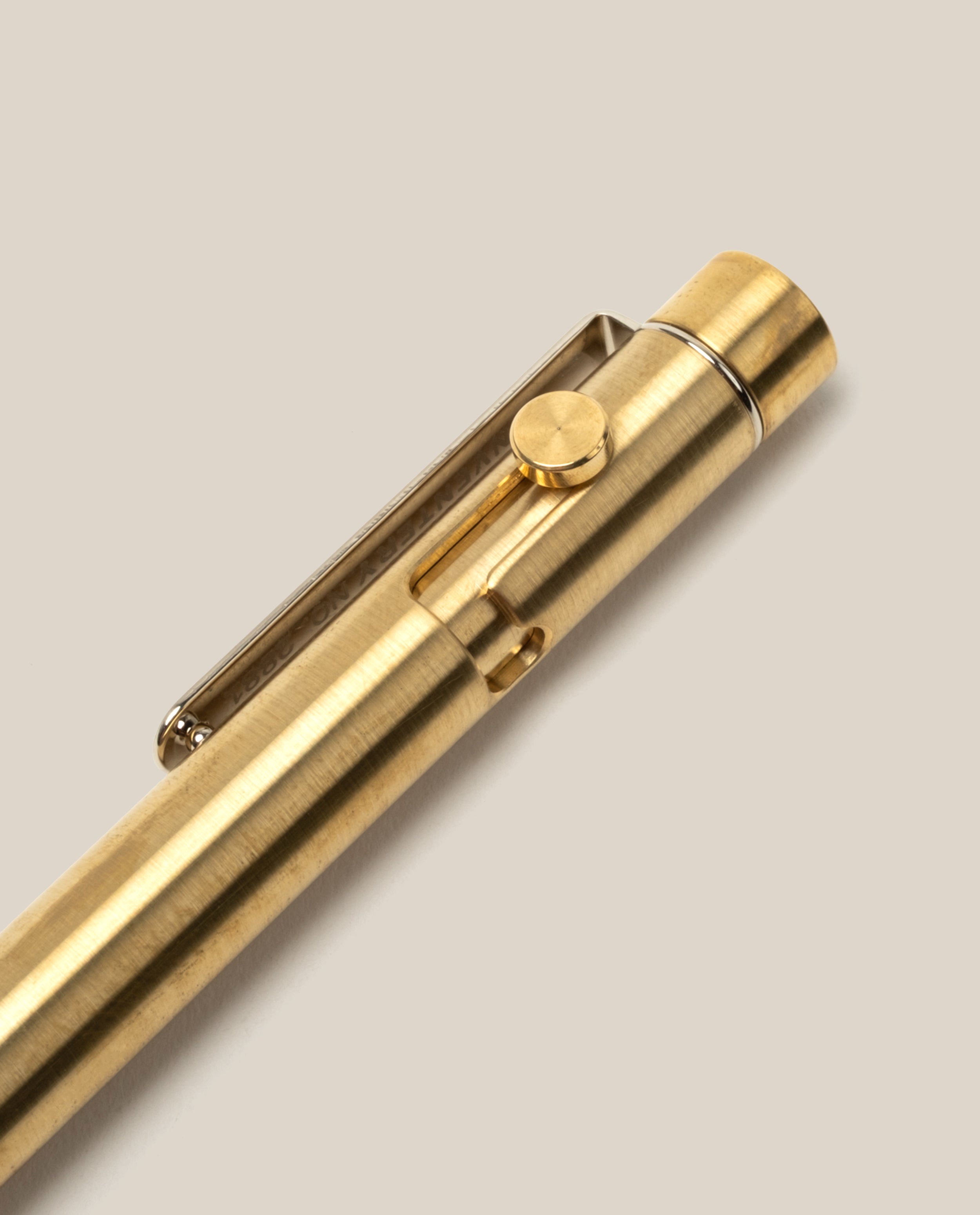 A close-up of the INVENTERY Bolt Action Pen (Brass), featuring a gold-colored solid brass body and a clip, photographed against a light beige background.