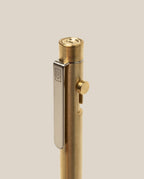 A close-up of the INVENTERY Bolt Action Pen (Brass), featuring a silver metal clip and a knurled button on top, set against a plain beige background, highlights the refined engineering of this solid brass writing instrument.