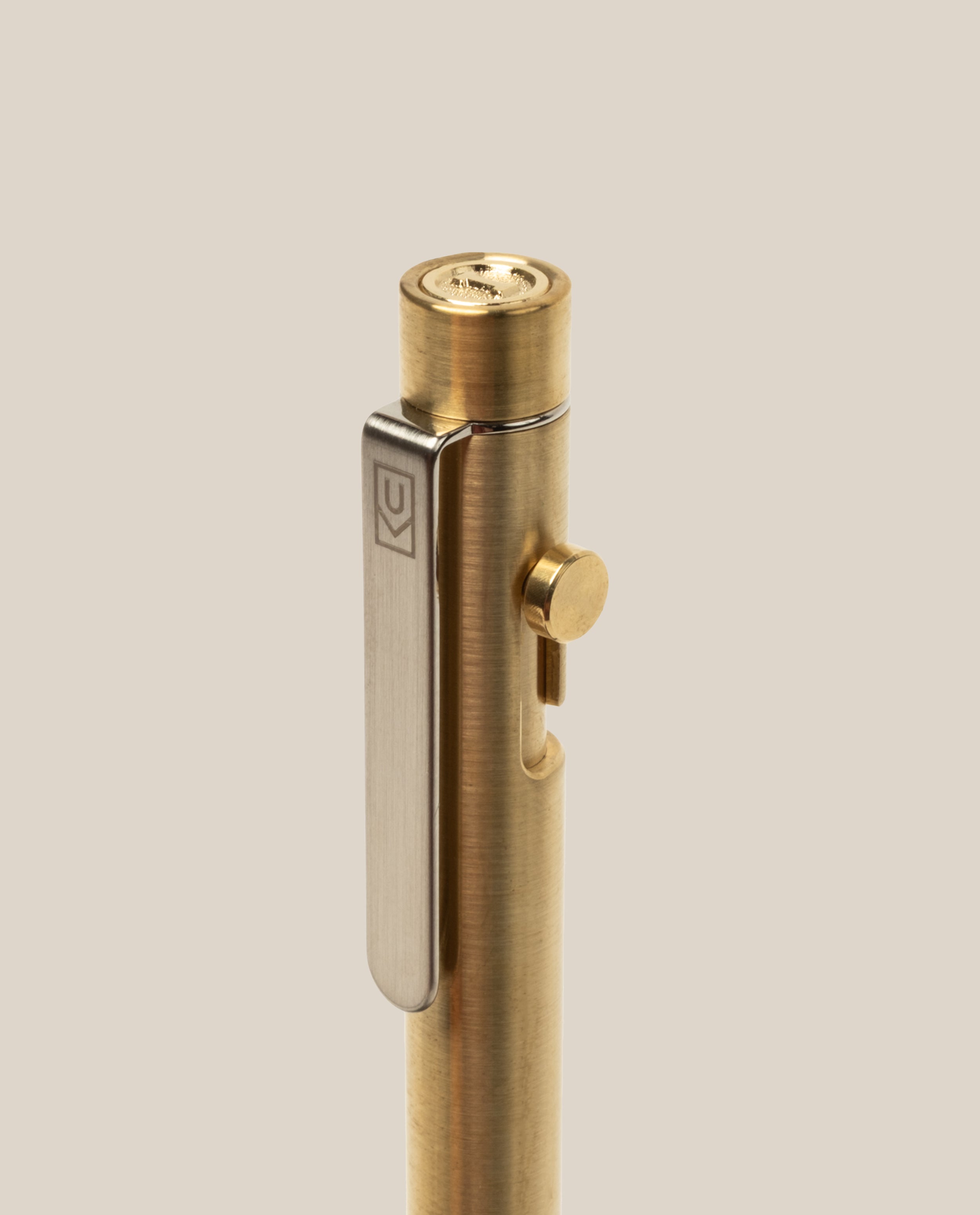 A close-up of the INVENTERY Bolt Action Pen (Brass), featuring a silver metal clip and a knurled button on top, set against a plain beige background, highlights the refined engineering of this solid brass writing instrument.