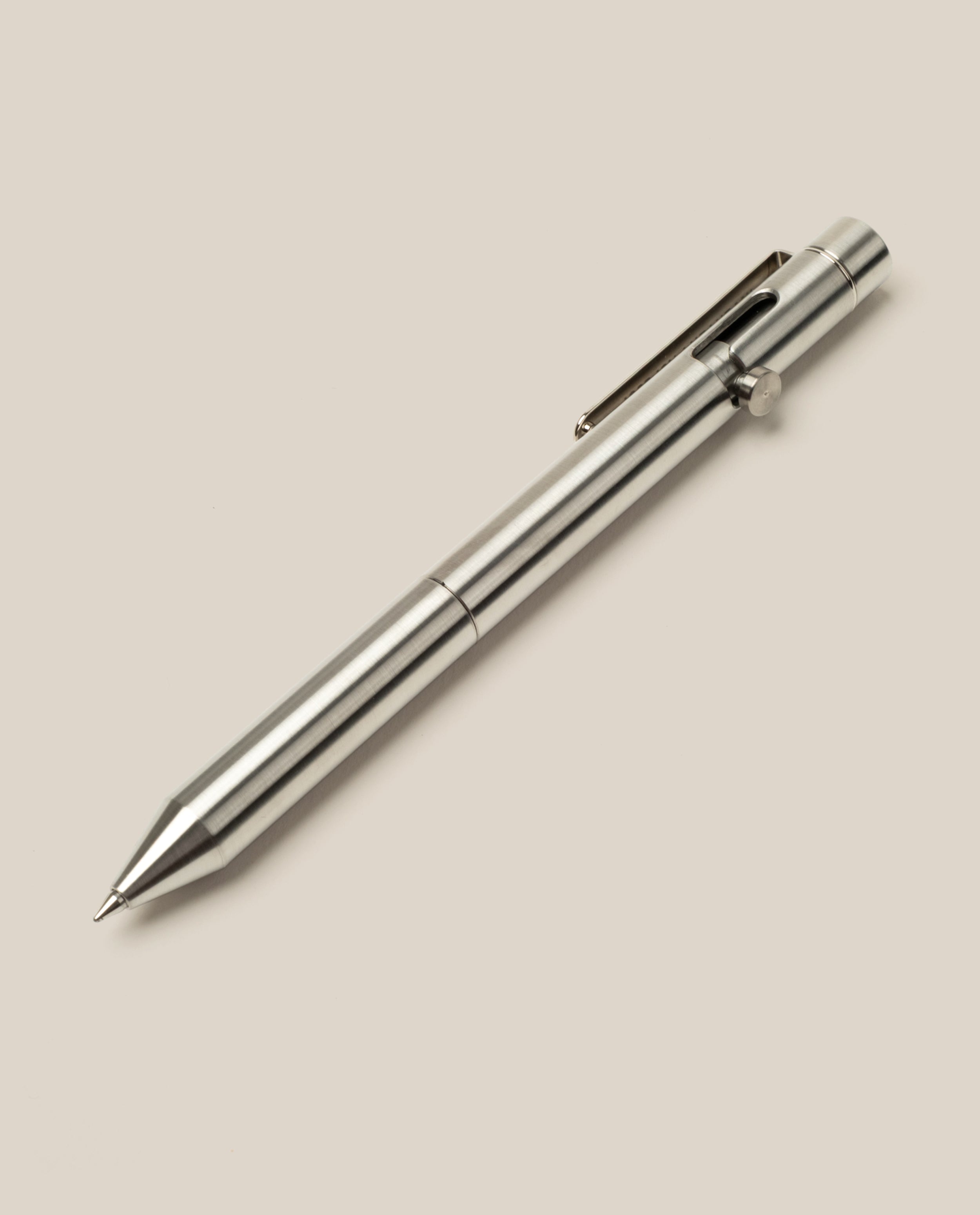 A sleek, metallic silver INVENTERY Bolt Action Pen (Stainless) with a clip and push-button mechanism is set diagonally from bottom left to top right, shown against a plain light beige background.
