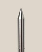 A close-up of the INVENTERY Bolt Action Pen (Stainless) with a brushed stainless steel finish, displayed vertically against a plain light beige background.
