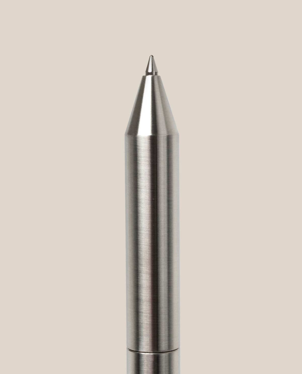 A close-up of the INVENTERY Bolt Action Pen (Stainless) with a brushed stainless steel finish, displayed vertically against a plain light beige background.