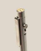 A close-up of the INVENTERY Bolt Action Pen (Stainless), featuring a gold-colored coin on top, a logo-engraved clip, and side button mechanism, set against a plain beige background.