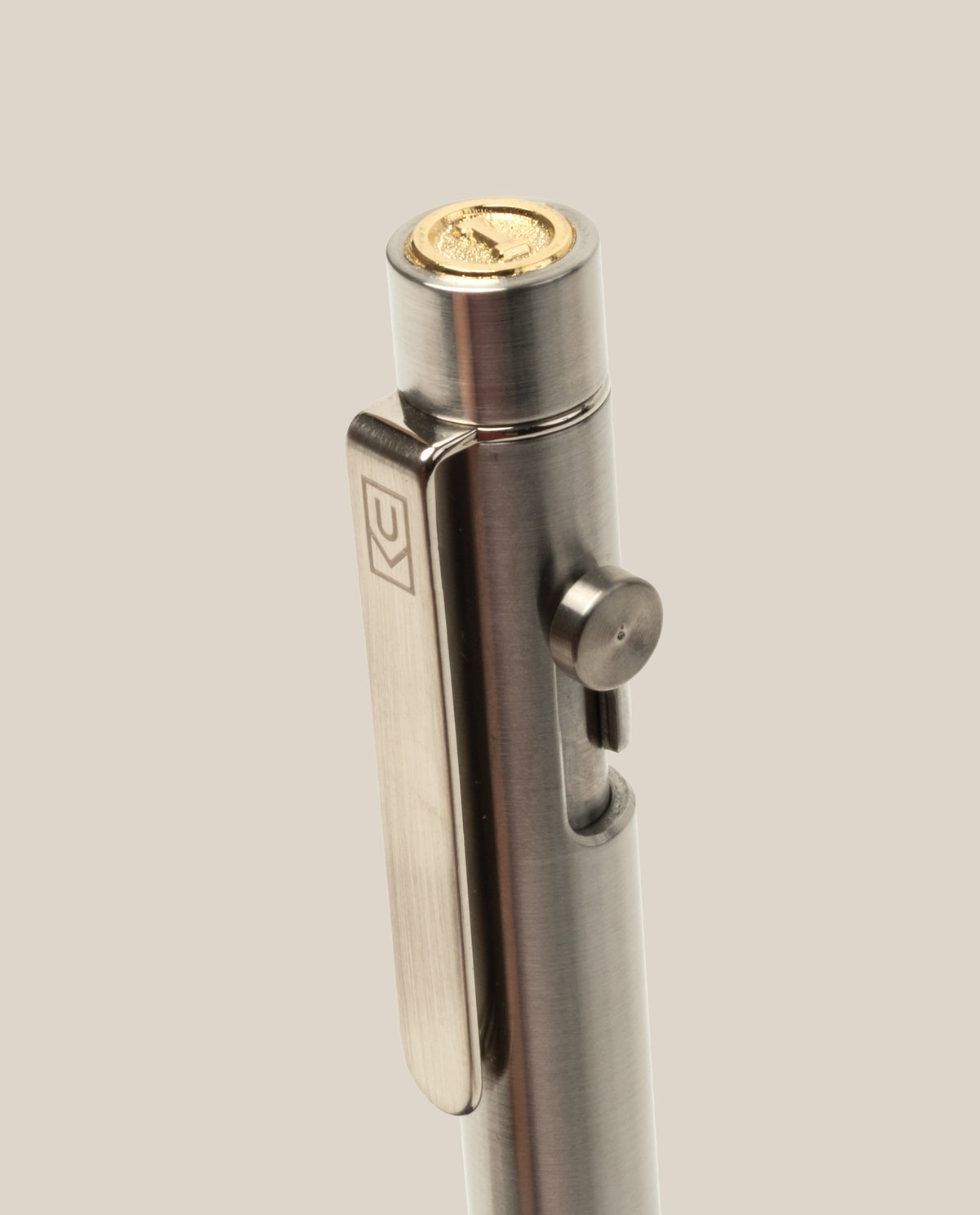 A close-up of the INVENTERY Bolt Action Pen (Stainless), featuring a gold-colored coin on top, a logo-engraved clip, and side button mechanism, set against a plain beige background.