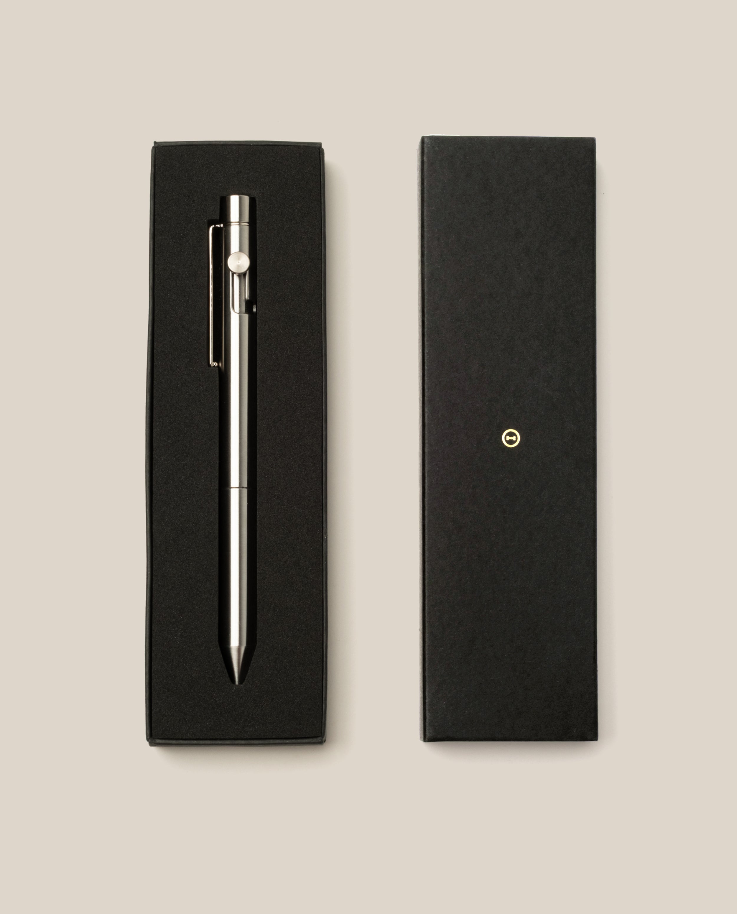 The INVENTERY Bolt Action Pen (Stainless) with a clip is displayed in an open black gift box next to its closed lid, which features a small gold emblem, all arranged on a light beige background.