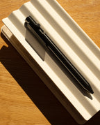 The INVENTERY Bolt Action Pen (Zirconium), a luxury writing instrument in black zirconium, rests atop a closed, textured white notebook as sunlight casts bold shadows across the wooden surface.