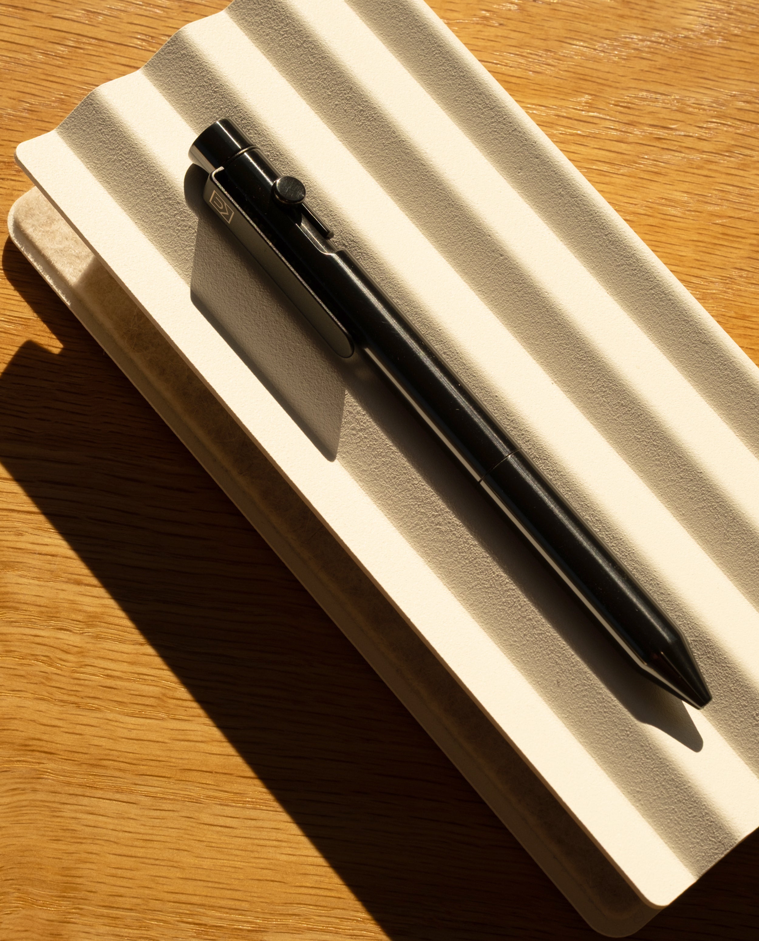 The INVENTERY Bolt Action Pen (Zirconium), a luxury writing instrument in black zirconium, rests atop a closed, textured white notebook as sunlight casts bold shadows across the wooden surface.