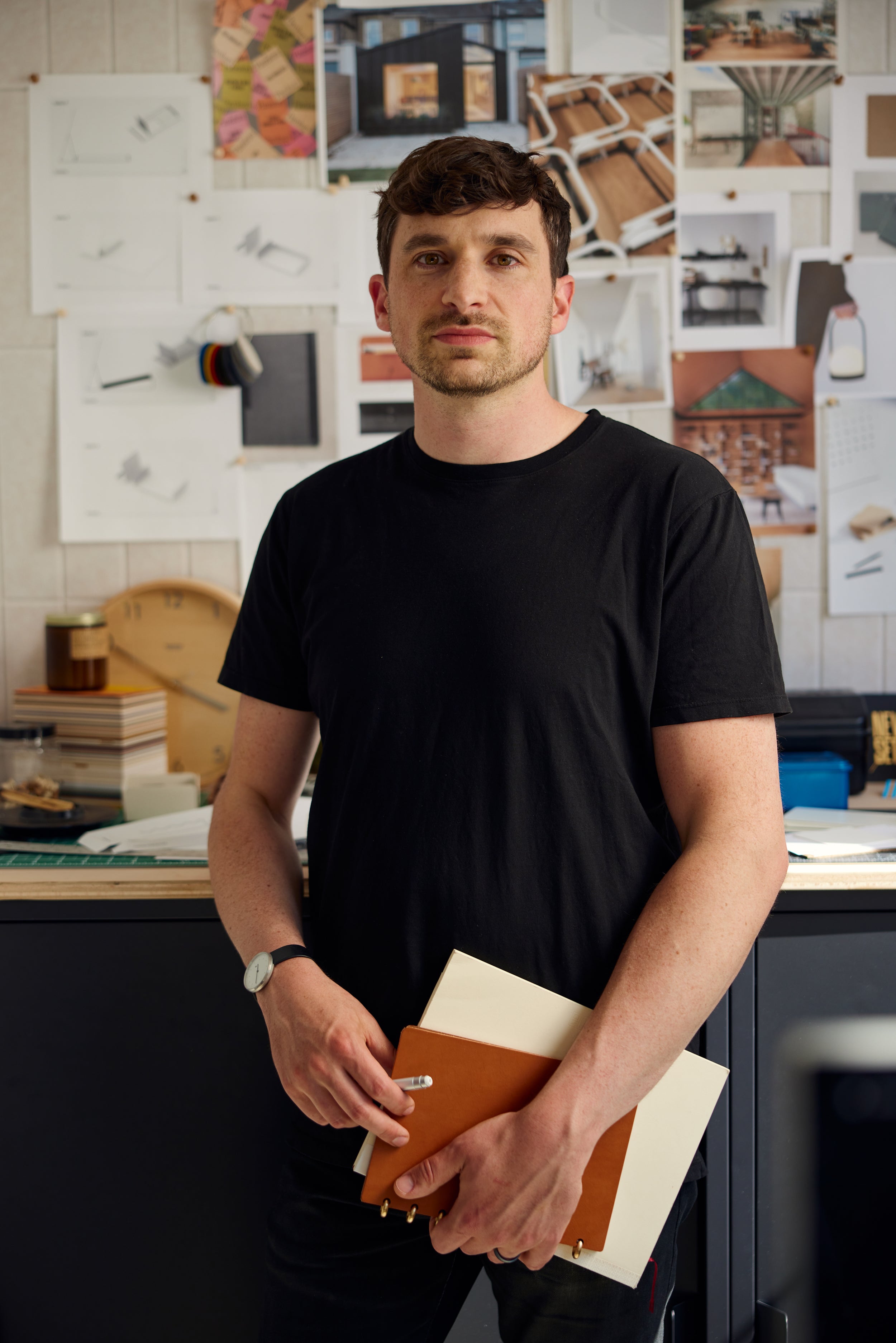 Man in a black t-shirt holding a notebook stands in an office with architectural sketches, photos, and models displayed on the wall behind him.