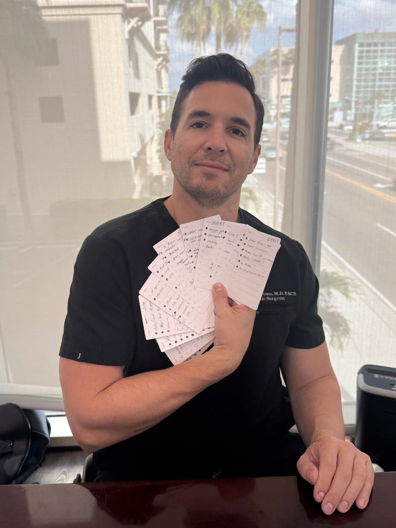 A man in black scrubs sits at a desk, smiling and holding several handwritten note cards fanned out in one hand. A city street and palm trees are visible through the window behind him.
